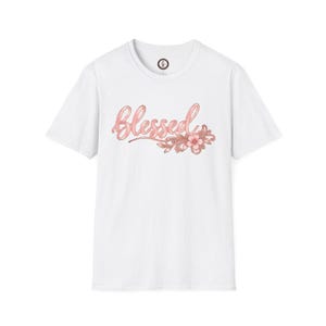 May include: A white short-sleeved t-shirt featuring the word Blessed in a metallic pink cursive script across the chest, accompanied by a decorative pink floral graphic with swirling vine details positioned to the right of the text.