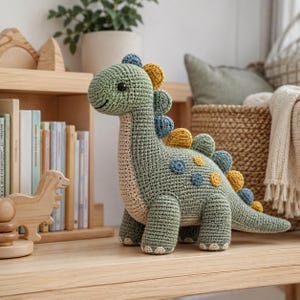 Può includere: Un giocattolo a forma di dinosauro realizzato all'uncinetto in verde salvia con la pancia color crema. Il dinosauro presenta piccole punte rotonde multicolori lungo la schiena e la coda nei toni del giallo, blu e verde, con occhi neri.