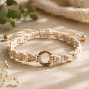 May include: A cream-colored macrame bracelet featuring a central circular wooden ring. The bracelet is constructed with intricate knotted cord and includes an adjustable sliding knot closure finished with small gold-toned metal beads at the ends.