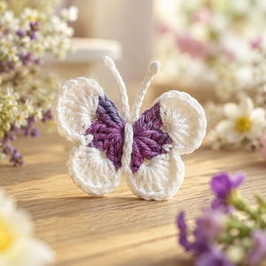 May include: A crocheted butterfly ornament featuring white outer wings and a purple and white variegated center body. The butterfly has two thin, upright white antennae and is constructed with a textured yarn stitch pattern throughout its form.