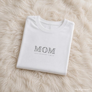 Könnte beinhalten: Ein weißes T-Shirt mit kurzen Ärmeln, das auf der Brust das Wort MOM in einer stilisierten schwarzen Serifenschrift zeigt. Unter dem Wort MOM ist der Satz LOVE WITHOUT LIMITS in einer kleineren, klaren schwarzen serifenlosen Schrift gedruckt.