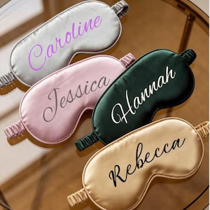 Peut inclure: Quatre masques de sommeil en satin avec des bandes élastiques en argent, rose, vert foncé et doré. Chaque masque présente un prénom personnalisé imprimé en police cursive : Caroline en violet, Jessica en gris, Hannah en blanc et Rebecca en noir.