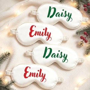 Könnte beinhalten: Vier weiße Schlafmasken aus Satin mit elastischen Bändern. Zwei Masken tragen den Namen Daisy in grüner Schreibschrift, zwei Masken tragen den Namen Emily in roter Schreibschrift. Die Masken sind auf einem weichen, hellen Stoff angeordnet.