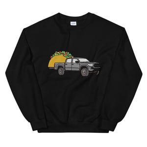 Puede incluir: Sudadera negra con un gráfico de una camioneta que lleva un taco grande. La camioneta es gris y el taco es amarillo con rellenos verdes y rojos.