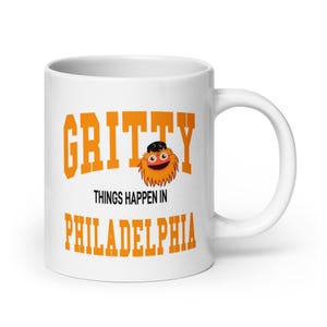 Puede incluir: Taza de cerámica blanca con una mascota de dibujos animados naranja y negra con el texto "Gritty Things Happen in Philadelphia" impreso en ella.