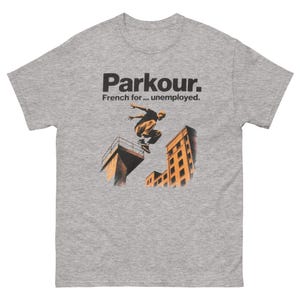 May include: A heather gray t-shirt featuring the word "Parkour" in black, with the phrase "French for... unemployed." The shirt has a graphic of a person doing parkour between buildings, in shades of brown and orange.