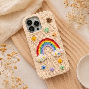 May include: A beige crochet phone case featuring a central rainbow motif with red, orange, yellow, green, blue, and purple stripes. The design includes two white clouds with small colorful accents and several small, textured star-shaped appliques.