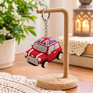 Può includere: Un portachiavi all'uncinetto a forma di auto rossa con un motivo della bandiera Union Jack sul tetto, finestrini grigi e ruote a bottone di colore chiaro, appeso a un anello e una catena in metallo argentato su un supporto rivestito in corda.