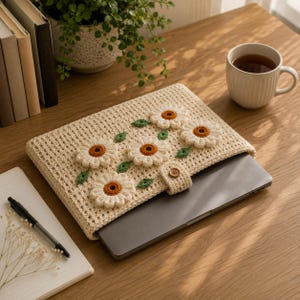 May include: A cream-colored crochet laptop sleeve featuring four dimensional white daisy appliques with brown centers and small green leaves. The sleeve has a matching crochet strap secured by a single brown wooden button, enclosing a dark gray laptop.
