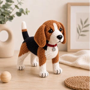 May include: A crocheted beagle dog figurine featuring a tricolor pattern of brown, black, and white yarn. The plush toy has long floppy ears, a black nose, and a red collar with a small, round, silver-colored metal tag hanging from the front.