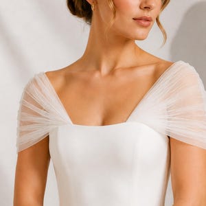 Può includere: Un abito bianco con scollo quadrato e maniche ad aletta in tulle plissettato trasparente. Il corpetto è realizzato in un tessuto bianco liscio e solido, offrendo un aspetto pulito e strutturato nella zona del petto e delle spalle.