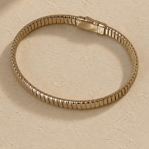 May include: A gold-toned metal bracelet featuring a flexible, ribbed, and segmented design. The bracelet includes a rectangular clasp mechanism with visible engravings that read AG925 and ITALY, indicating its metallic composition and origin.
