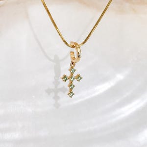 Puede incluir: Un collar de cadena de serpiente dorada con una peque&ntilde;a anilla circular dorada de la que cuelga un colgante en forma de cruz. La cruz est&aacute; engastada con varias piedras preciosas peque&ntilde;as de color verde claro y corte redondo en sus brazos.