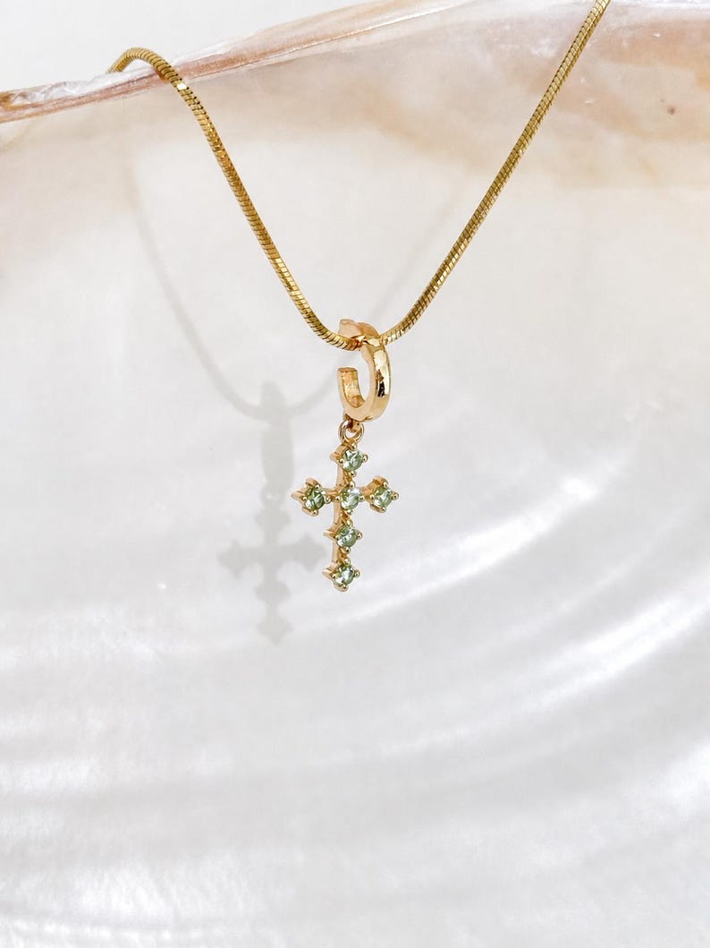 Puede incluir: Un collar de cadena de serpiente dorada con una peque&ntilde;a anilla circular dorada de la que cuelga un colgante en forma de cruz. La cruz est&aacute; engastada con varias piedras preciosas peque&ntilde;as de color verde claro y corte redondo en sus brazos.
