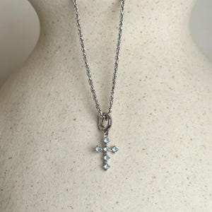 Puede incluir: Un collar de cadena de cuerda en tono plateado con un peque&ntilde;o colgante de cruz. La cruz est&aacute; engastada con cinco piedras de color azul claro de corte cuadrado dispuestas en un patr&oacute;n vertical y horizontal, suspendida de una argolla circular.