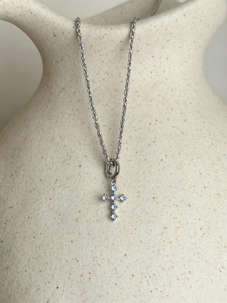 Puede incluir: Un collar de cadena de cuerda en tono plateado con un peque&ntilde;o colgante de cruz. La cruz est&aacute; engastada con cinco piedras de color azul claro de corte cuadrado dispuestas en un patr&oacute;n vertical y horizontal, suspendida de una argolla circular.