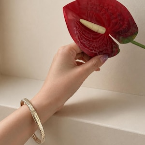 May include: A gold-toned bracelet featuring a ribbed, textured surface and a smooth, rectangular clasp mechanism. The bracelet is worn on the wrist, showcasing a metallic finish with consistent vertical ridges along the entire length of the band.