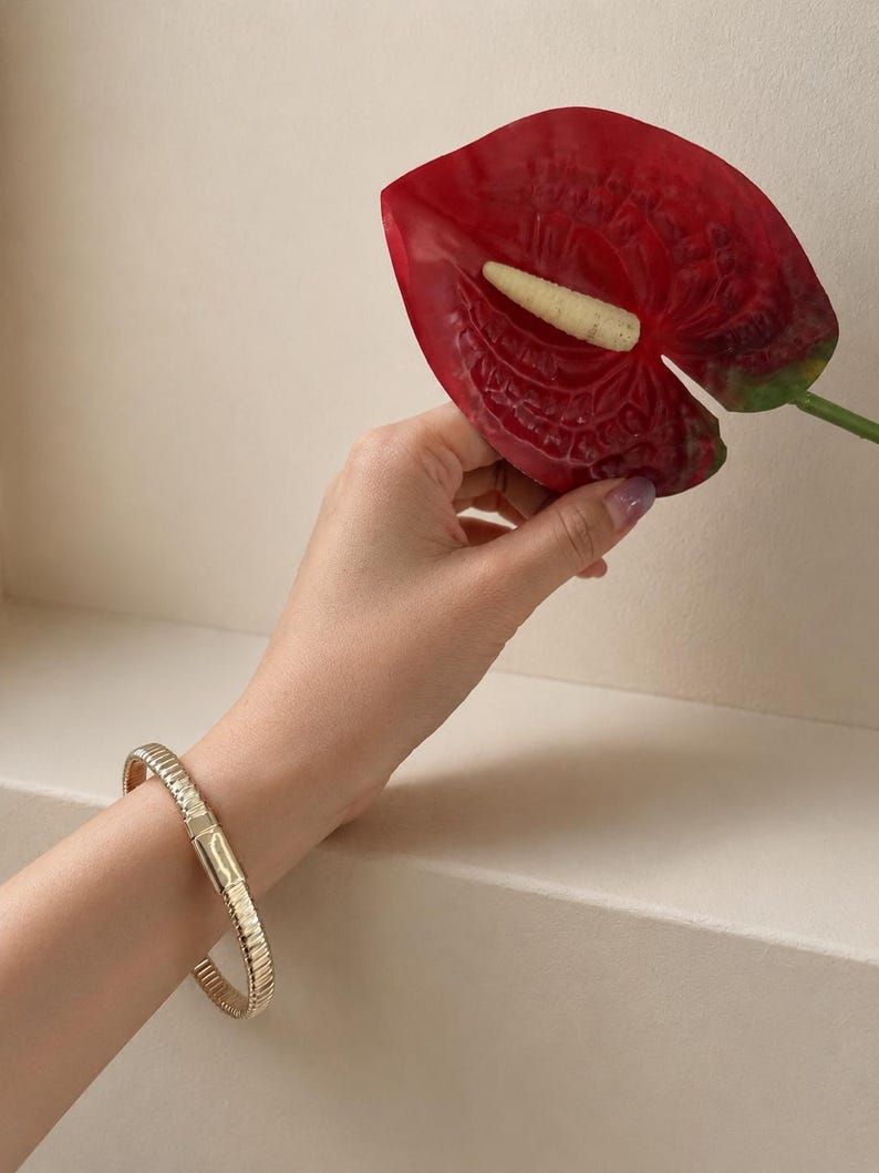May include: A gold-toned bracelet featuring a ribbed, textured surface and a smooth, rectangular clasp mechanism. The bracelet is worn on the wrist, showcasing a metallic finish with consistent vertical ridges along the entire length of the band.