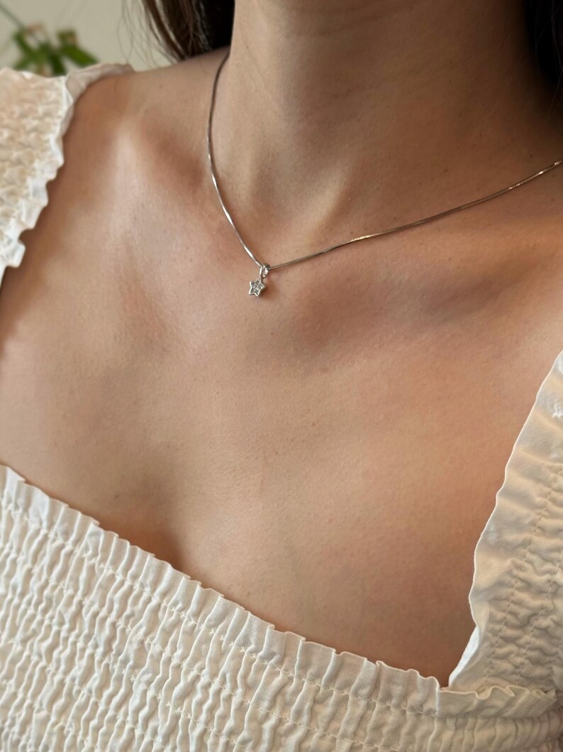 Op de afbeelding: Een zilverkleurige ketting met een kleine, vijfpuntige sterhanger. De sterhanger is in het midden versierd met een enkele, heldere, fonkelende steen. De ketting rust op het sleutelbeen en heeft een verfijnd schakelontwerp.