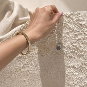 May include: A gold-toned bangle bracelet featuring a textured, ribbed surface design. The bracelet is circular in shape and has a polished metallic finish that reflects light, creating a consistent pattern of vertical lines around the entire band.