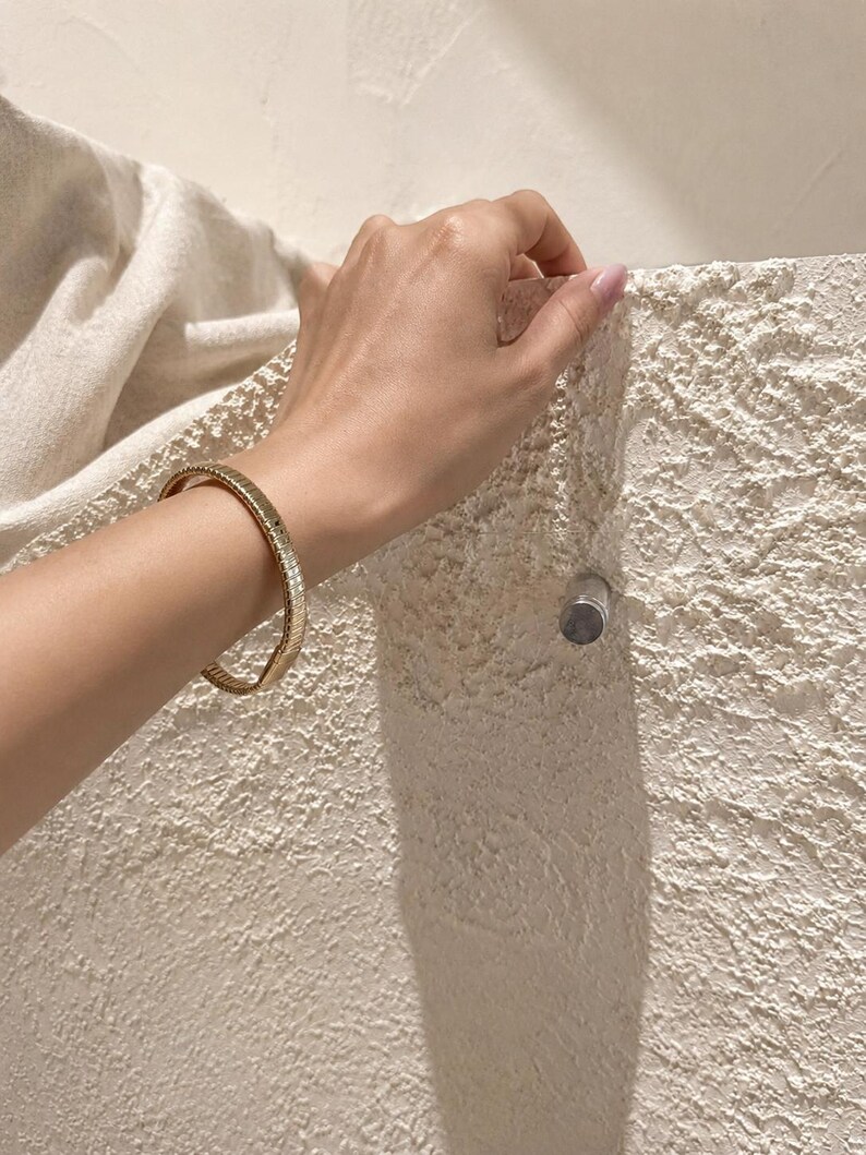 May include: A gold-toned bangle bracelet featuring a textured, ribbed surface design. The bracelet is circular in shape and has a polished metallic finish that reflects light, creating a consistent pattern of vertical lines around the entire band.
