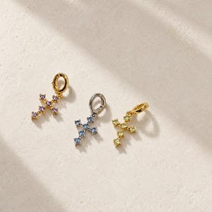 Puede incluir: Tres dijes en forma de cruz, cada uno con una peque&ntilde;a argolla circular en la parte superior. El primer dije es de tono dorado con piedras moradas, el segundo es de tono plateado con piedras azules y el tercero es de tono dorado con piedras amarillas.