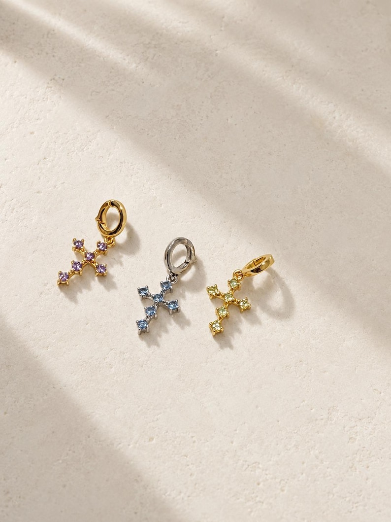 Puede incluir: Tres dijes en forma de cruz, cada uno con una peque&ntilde;a argolla circular en la parte superior. El primer dije es de tono dorado con piedras moradas, el segundo es de tono plateado con piedras azules y el tercero es de tono dorado con piedras amarillas.