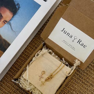 Puede incluir: Un collar de cadena en tono dorado y un dije de cruz en tono dorado por separado, ambos con peque&ntilde;as piedras incrustadas, sobre una bolsa de tela beige dentro de una caja de regalo de cart&oacute;n marr&oacute;n con la etiqueta Juna Rae y Clip.Collect.Create.