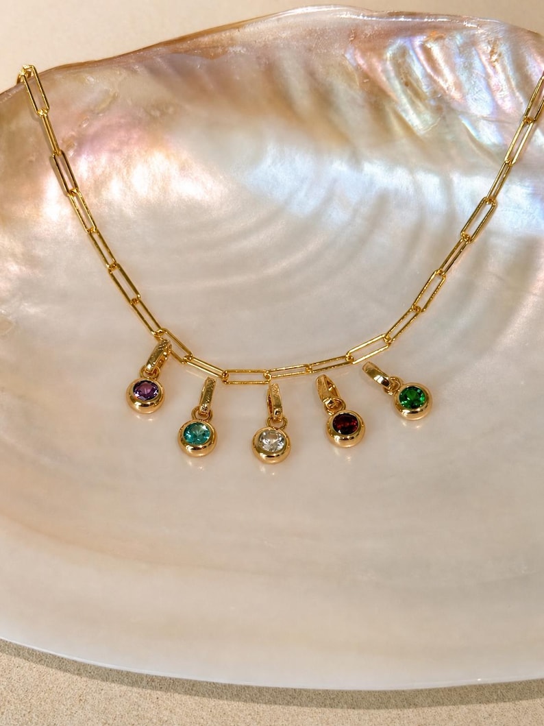Puede incluir: Un collar de cadena dorada con eslabones tipo clip y cinco dijes colgantes, cada uno con una piedra preciosa redonda engastada en bisel de color morado, turquesa, transparente, rojo oscuro y verde.