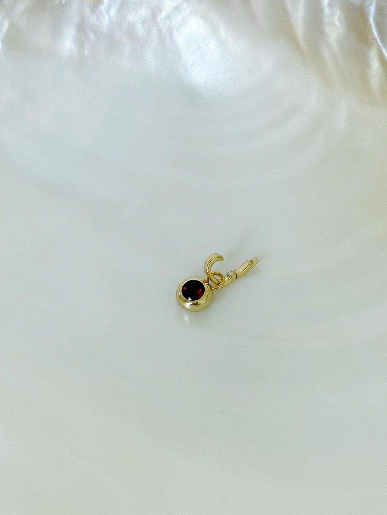 Puede incluir: Un peque&ntilde;o dije de oro con una piedra preciosa redonda de color rojo oscuro engastada en un bisel de oro pulido. El dije incluye una peque&ntilde;a argolla circular unida a un mecanismo de cierre con bisagra para engancharlo a una cadena.