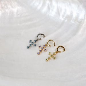 Puede incluir: Tres pendientes de aro con colgantes en forma de cruz. El primero es plateado con piedras azules, el segundo es dorado con piedras moradas y el tercero es dorado con piedras verde claro. Cada cruz cuenta con cinco peque&ntilde;as piedras incrustadas.