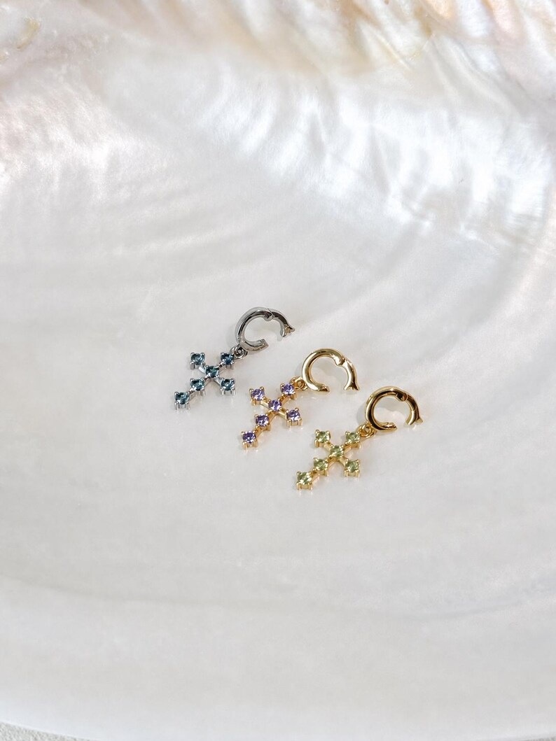 Puede incluir: Tres pendientes de aro con colgantes en forma de cruz. El primero es plateado con piedras azules, el segundo es dorado con piedras moradas y el tercero es dorado con piedras verde claro. Cada cruz cuenta con cinco peque&ntilde;as piedras incrustadas.