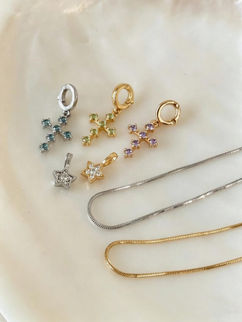 Puede incluir: Tres dijes de cruz con piedras azules, verdes y moradas, dos dijes de estrella con piedras transparentes y dos cadenas de serpiente en tonos plata y oro. Los dijes de cruz tienen argollas circulares para sujetarlos a las cadenas.