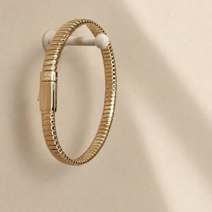 May include: A gold-toned bracelet featuring a flexible, ribbed, and segmented band design. The piece includes a rectangular, smooth-surfaced clasp mechanism that is integrated into the circular structure of the jewelry item.
