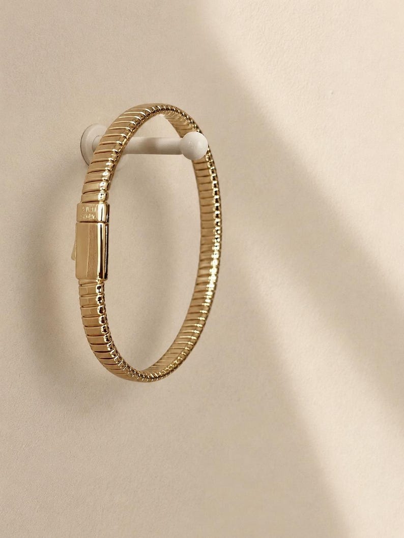 May include: A gold-toned bracelet featuring a flexible, ribbed, and segmented band design. The piece includes a rectangular, smooth-surfaced clasp mechanism that is integrated into the circular structure of the jewelry item.
