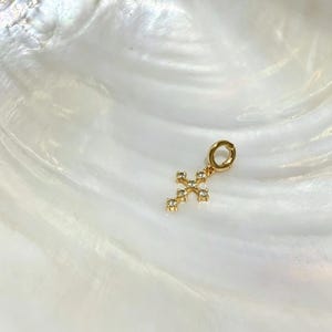 Puede incluir: Un colgante en forma de cruz de tono dorado con peque&ntilde;as piedras claras y brillantes engastadas a lo largo de la estructura. El colgante incluye una anilla circular en la parte superior para su sujeci&oacute;n, con un acabado met&aacute;lico pulido y dise&ntilde;o.