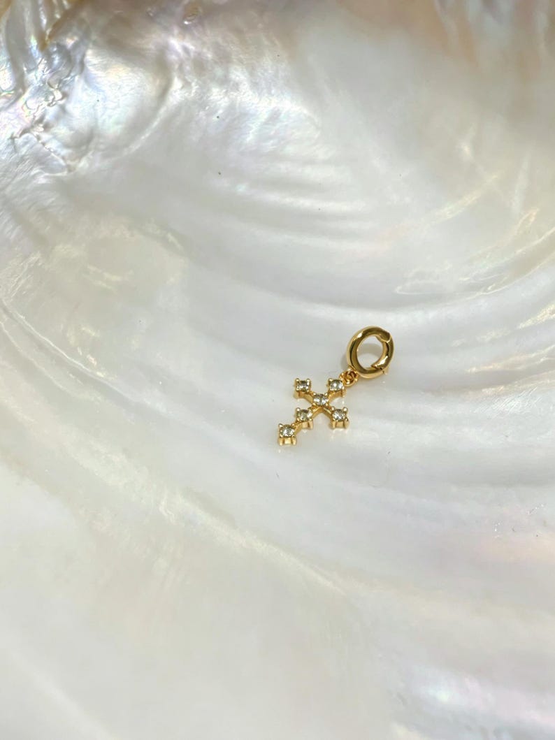 Puede incluir: Un colgante en forma de cruz de tono dorado con peque&ntilde;as piedras claras y brillantes engastadas a lo largo de la estructura. El colgante incluye una anilla circular en la parte superior para su sujeci&oacute;n, con un acabado met&aacute;lico pulido y dise&ntilde;o.