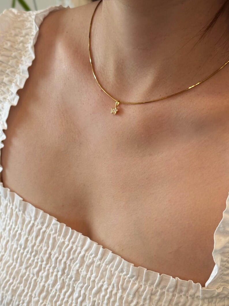Op de afbeelding: Een goudkleurige ketting met een kleine, vijfpuntige sterhanger. De hanger is in het midden van de ketting geplaatst en bevat een enkele, kleine, heldere, sprankelende steen in het midden. De ketting rust op een wit kledingstuk van textuurrijke stof.