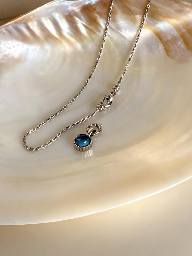 Pode incluir: Um colar de corrente de corda prateada com um pequeno pendente redondo que apresenta uma pedra caboch&atilde;o azul profunda e lisa, engastada numa moldura de metal estriado. O colar inclui um fecho de lagosta e um pequeno anel de liga&ccedil;&atilde;o.