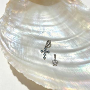 Puede incluir: Dos dijes de metal en tono plateado. Uno tiene forma de cruz con peque&ntilde;as piedras azul claro y un anillo de uni&oacute;n. El segundo dije tiene forma de estrella con peque&ntilde;as piedras transparentes y cuenta con una peque&ntilde;a argolla para colgar.