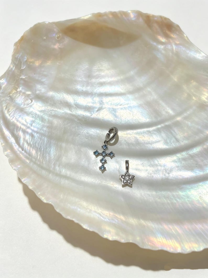 Puede incluir: Dos dijes de metal en tono plateado. Uno tiene forma de cruz con peque&ntilde;as piedras azul claro y un anillo de uni&oacute;n. El segundo dije tiene forma de estrella con peque&ntilde;as piedras transparentes y cuenta con una peque&ntilde;a argolla para colgar.