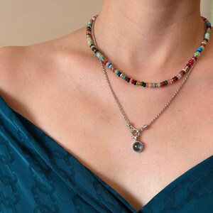 Pode incluir: Um conjunto de colares em camadas composto por um fio curto de pequenas contas multicoloridas em tons de vermelho, verde, azul e dourado, e uma corrente prateada mais longa com um pendente circular com uma pedra escura e iridescente.