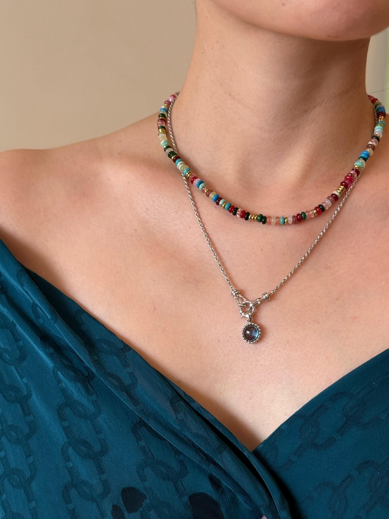 Pode incluir: Um conjunto de colares em camadas composto por um fio curto de pequenas contas multicoloridas em tons de vermelho, verde, azul e dourado, e uma corrente prateada mais longa com um pendente circular com uma pedra escura e iridescente.
