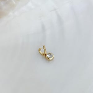 Puede incluir: Un peque&ntilde;o dije de oro con una piedra redonda, transparente y facetada, engastada en un bisel de oro pulido. El dije incluye una peque&ntilde;a argolla de oro abierta en la parte superior, dise&ntilde;ada para sujetarse a una cadena o una pulsera.