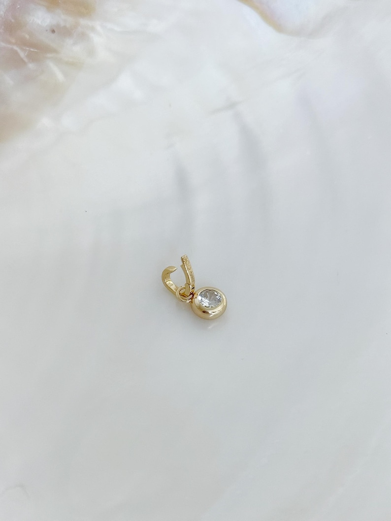 Puede incluir: Un peque&ntilde;o dije de oro con una piedra redonda, transparente y facetada, engastada en un bisel de oro pulido. El dije incluye una peque&ntilde;a argolla de oro abierta en la parte superior, dise&ntilde;ada para sujetarse a una cadena o una pulsera.
