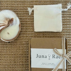 Puede incluir: Una vela en concha de nautilo, una peque&ntilde;a bolsa de tela color hueso con cord&oacute;n y una caja de regalo cuadrada de cart&oacute;n marr&oacute;n atada con una cinta beige. La caja tiene una etiqueta blanca con el texto Juna Rae y WEAR IT YOUR WAY CLIP.COLLECT.CREATE.