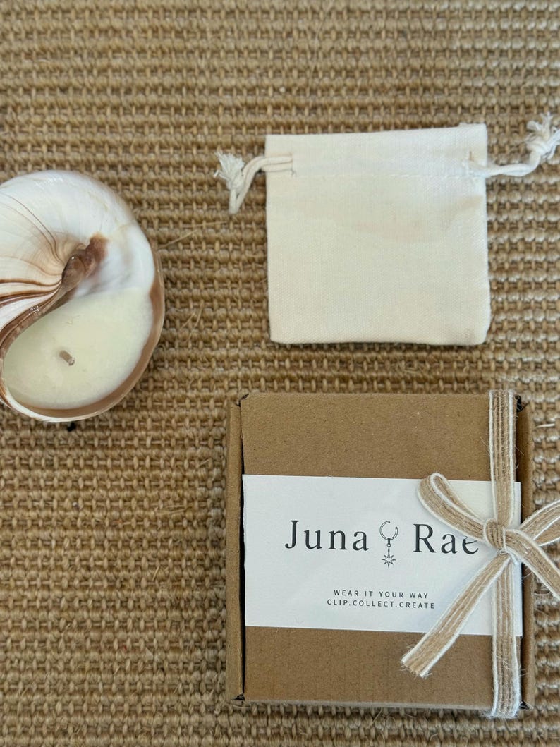Puede incluir: Una vela en concha de nautilo, una peque&ntilde;a bolsa de tela color hueso con cord&oacute;n y una caja de regalo cuadrada de cart&oacute;n marr&oacute;n atada con una cinta beige. La caja tiene una etiqueta blanca con el texto Juna Rae y WEAR IT YOUR WAY CLIP.COLLECT.CREATE.