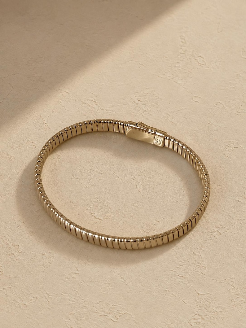 May include: A gold-toned metal bracelet featuring a flexible, ribbed, and segmented design. The bracelet includes a rectangular clasp mechanism with visible engravings that read AG925 and ITALY, indicating its metallic composition and origin.