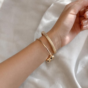 May include: Two gold-toned bracelets worn on the wrist. One bracelet features a textured, ribbed surface with a uniform pattern. The second bracelet is a smooth, open-ended cuff design finished with a polished spherical bead at each end.