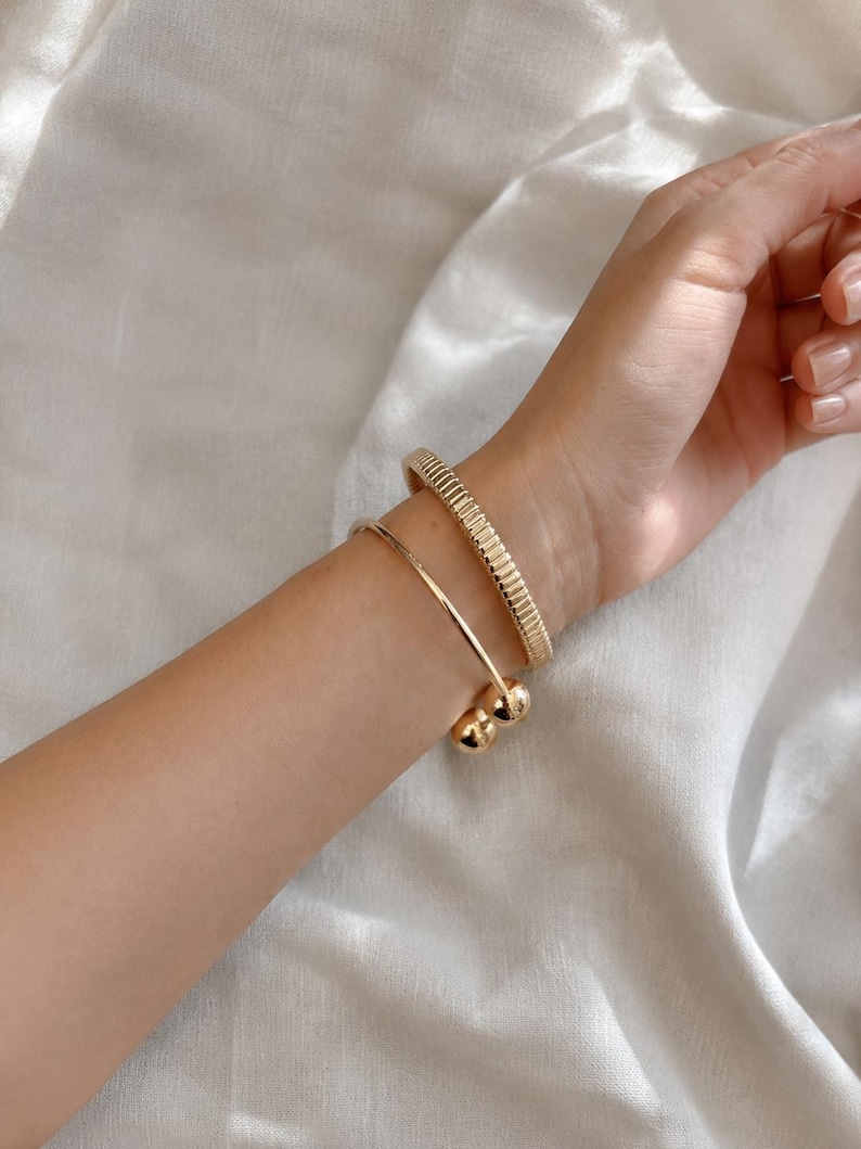 May include: Two gold-toned bracelets worn on the wrist. One bracelet features a textured, ribbed surface with a uniform pattern. The second bracelet is a smooth, open-ended cuff design finished with a polished spherical bead at each end.